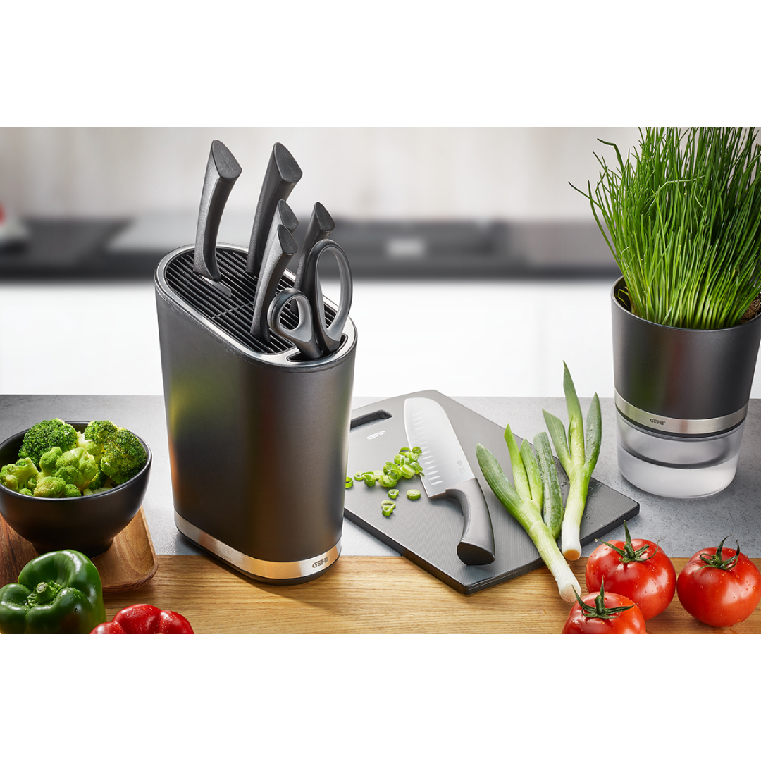 Virtuves galda virsma ar garšaugu podu, griešanas dēlīti un santoku nazi. Blakus novietots nažu turētājs gefu Smartilne ar 5 dažādu veidu nažiem un šķērēm. Ar Gefu santoku nazi uz pelēka Gefu dēlīša griež zaļus sīpollokus.